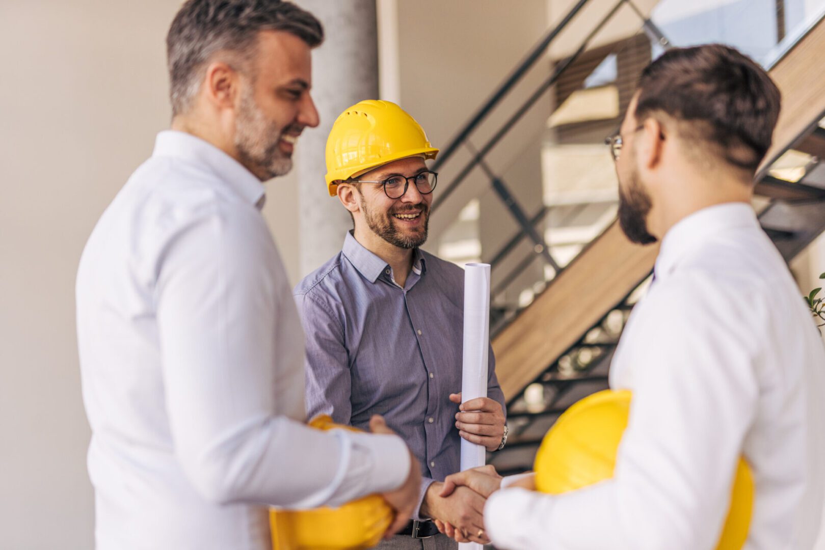 Three architects shaking hands on a construction site after finalizing an agreement, one holding blueprints, embodying teamwork and collaboration in a successful project